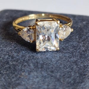 Peut inclure: Une bague en or avec une grande pierre précieuse rectangulaire et transparente au centre, flanquée de deux pierres en forme de poire plus petites. L'anneau est en or poli.