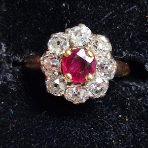 Peut inclure: Une bague vintage avec un rubis central facetté, entouré d'un halo de diamants clairs. La monture de la bague est en or, sur fond sombre.