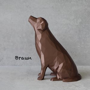 May include: A brown geometric dog sculpture in a sitting position, with its head tilted upwards. The word "Braun" is written in black script on the lower left. The sculpture is on a light gray surface against a matching background.