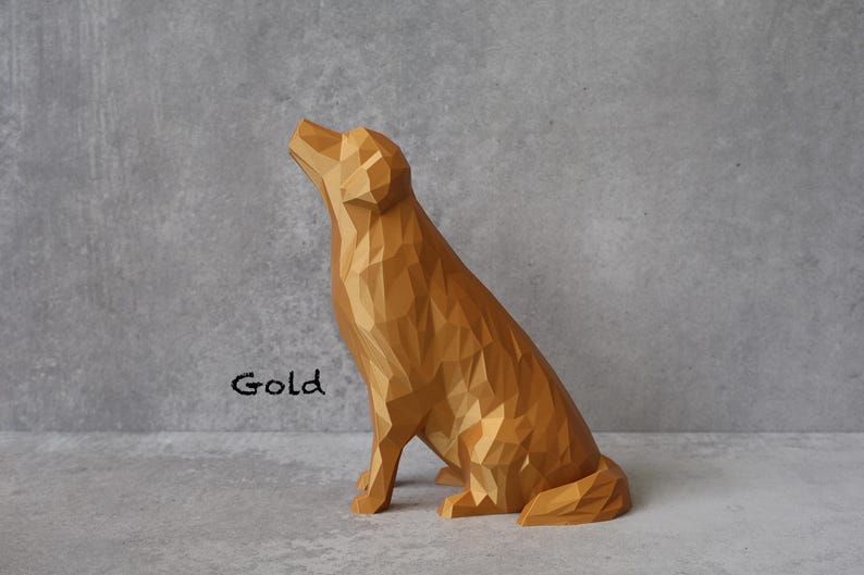 May include: A gold-coloured, geometric dog sculpture in a seated position, with its head tilted upwards. The word "Gold" is written in black script on the left side of the sculpture. The dog has a faceted design, giving it a modern look.