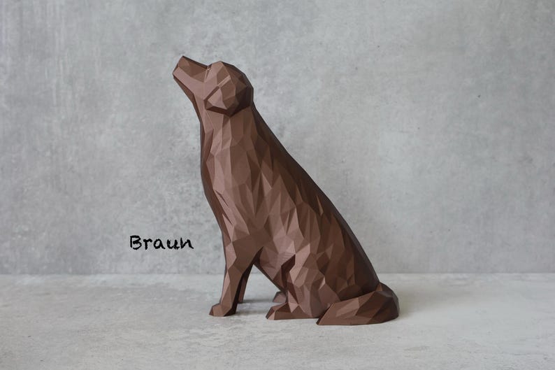 May include: A brown geometric dog sculpture in a seated position, with its head tilted upwards. The sculpture has a faceted design and is set against a grey background. The word "Braun" is written in black cursive.