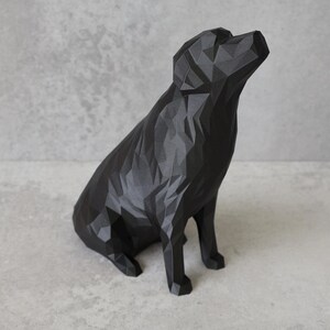 May include: A black geometric dog sculpture with a faceted design. The dog is seated, head tilted upwards. The sculpture is made of matte black material and sits on a light grey surface. A modern decorative piece.