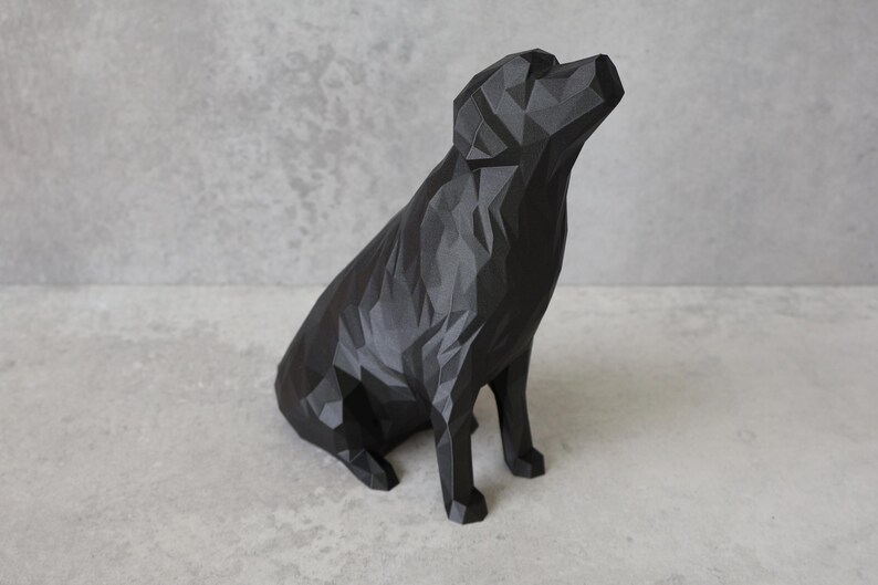May include: A black geometric dog sculpture with a faceted design. The dog is seated, head tilted upwards. The sculpture is made of matte black material and sits on a light grey surface. A modern decorative piece.