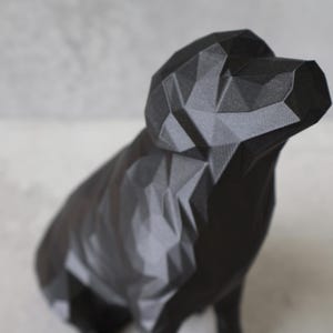 May include: A black geometric dog sculpture with a faceted design. The dog is in a sitting position, with its head slightly raised. The sculpture is made of a dark material and has a matte finish. The background is a light grey.