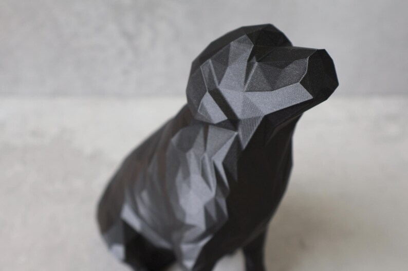 May include: A black geometric dog sculpture with a faceted design. The dog is in a sitting position, with its head slightly raised. The sculpture is made of a dark material and has a matte finish. The background is a light grey.
