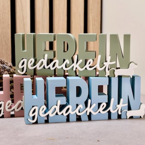May include: Two sets of stacked wooden letters spelling "HEREIN gedackelt" in sage green and light blue. Each set features white cursive text and a small white dachshund silhouette attached to the right side of the base.