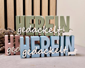 May include: Two sets of stacked wooden letters spelling "HEREIN gedackelt" in sage green and light blue. Each set features white cursive text and a small white dachshund silhouette attached to the right side of the base.