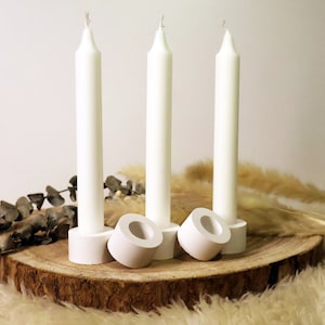 May include: Three white pillar candles on a wooden base with three white candle holders. The candles are on a white fluffy surface with dried eucalyptus branches and dried pampas grass.