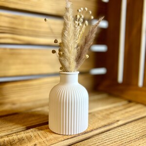 May include: A white ceramic vase with vertical ridges, holding a bouquet of dried pampas grass and other dried flowers.