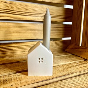 May include: A white ceramic house-shaped candle holder with a gray candle inside. The holder is sitting on a wooden surface.