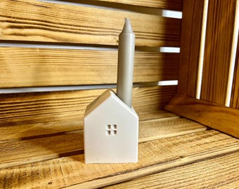 May include: A white ceramic house-shaped candle holder with a grey candle inside. The holder is sitting on a wooden surface.