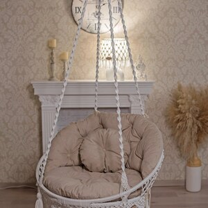 May include: White hanging chair with a beige cushion and fringe trim. The chair is suspended from the ceiling with white ropes.