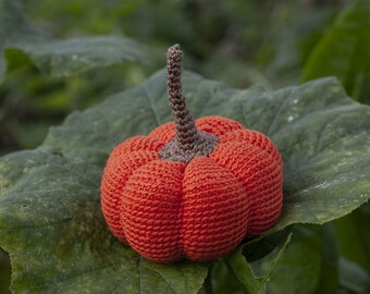 May include: A small, round, orange crocheted pumpkin featuring a textured, ribbed surface and a central, upright brown crocheted stem. The pumpkin is crafted with a consistent yarn stitch pattern, creating a soft, rounded shape with distinct segments.
