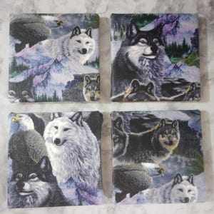 May include: Set of four square coasters featuring wolf and eagle imagery. Each coaster displays a different wolf portrait against a backdrop of mountains and trees. The color palette includes shades of gray, white, and purple.