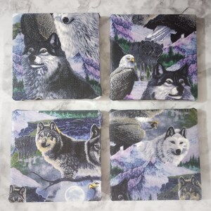 May include: Set of four square coasters featuring wildlife scenes. Each coaster displays a different image of wolves and eagles in a mountain landscape with shades of purple, blue, and green. The coasters are likely made of a durable material.