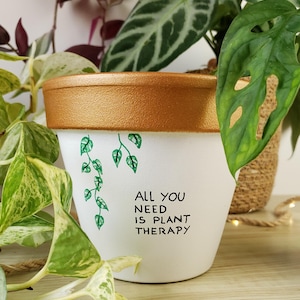 Può includere: Vaso per piante in ceramica bianca con bordo dorato e la scritta "ALL YOU NEED IS PLANT THERAPY" in nero. Foglie e viti verdi sono dipinte sul vaso. Il vaso è circondato da piante verdi.