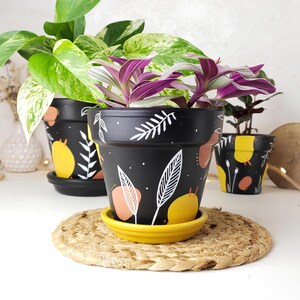 Op de afbeelding: Drie zwarte keramieken plantenpotten met bijpassende schotels, voorzien van handgeschilderde witte bladvormen, gele cirkels en terracotta-kleurige vormen. De potten bevatten diverse groene en paars gestreepte kamerplanten op een mat.