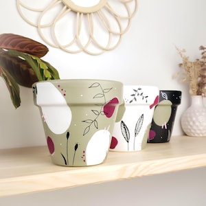 Peut inclure: Trois pots de fleurs en céramique peints à la main avec des motifs floraux. Les pots sont dans des tons de vert, blanc et noir, avec des accents bordeaux. Les pots sont disposés sur une étagère en bois, avec un miroir et une plante en arrière-plan.