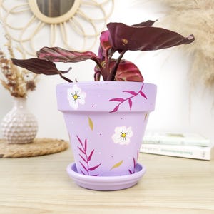 Peut inclure: Un pot de fleur en céramique de couleur lavande avec une soucoupe assortie, décoré de fleurs blanches, d'accents jaunes et de feuilles violettes. Le pot contient une plante aux feuilles bordeaux foncé. Le pot est posé sur une surface en bois.
