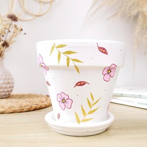 Peut inclure: Pot de fleur en céramique blanche avec soucoupe assortie, orné de motifs floraux roses et de feuilles dorées. Le pot est cylindrique avec un dessus légèrement évasé. Un ajout charmant pour les plantes.
