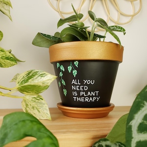 Peut inclure: Pot de plante noir avec un bord et une soucoupe dorés, portant l'inscription blanche "ALL YOU NEED IS PLANT THERAPY". La plante verte est dans le pot. Des feuilles vertes décoratives sont peintes.