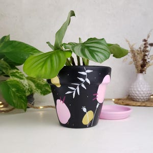 Peut inclure: Un pot de plante en céramique noire avec un bord rose et une soucoupe assortie. Le pot est décoré de motifs floraux abstraits blancs, roses et jaunes. Une plante verte à grandes feuilles est plantée à l'intérieur.