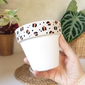 Peut inclure: Un pot de fleurs en céramique blanche doté d'un large rebord orné d'un motif léopard répétitif en noir et cuivre métallique. Le corps principal du pot est d'un blanc mat uni, offrant un contraste net avec la bordure à motif animalier.