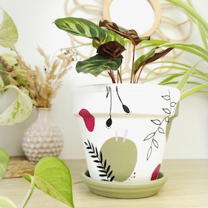 Può includere: Un vaso di fiori in ceramica bianca dipinta con un piattino verde, con disegni botanici astratti in nero, rosa e verde. Il vaso contiene una pianta da interno con foglie verdi e bordeaux. Il vaso è su una superficie di legno.