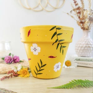 Puede incluir: Una maceta amarilla pintada con un diseño floral. La maceta presenta flores blancas, hojas verdes y detalles en rojo. La maceta está sobre una superficie de madera con otros elementos decorativos en el fondo.