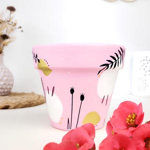 Peut inclure: Un pot de fleurs peint en rose avec un design fantaisiste. Le pot présente des formes florales et abstraites blanches, dorées et noires. Le pot est entouré de fleurs roses.