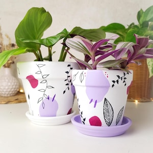 Op de afbeelding: Twee witte keramische bloempotten met handgeschilderde ontwerpen. De potten hebben paarse en roze abstracte vormen, zwarte bladeren en ranken. De potten zijn gevuld met groene en paarse planten en staan op bijpassende schotels.