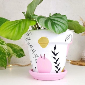 Peut inclure: Pot de plante en céramique blanche avec soucoupe rose, décoré d'art au trait noir, d'accents dorés et de formes roses. Le pot contient une plante verte avec de grandes feuilles veinées. Le design est fantaisiste et moderne.