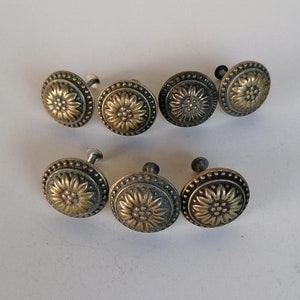 Op de afbeelding: Acht antieke goudkleurige kastknoppen met een bloemmotief. Elke knop heeft een kleine schroef om aan een kastdeur te bevestigen.