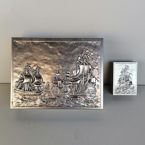 Op de afbeelding: Een zilverkleurige rechthoekige doos en een kleinere rechthoekige doos, beide met een reliëfontwerp van zeilschepen. De grotere doos heeft een gedetailleerde scène van meerdere schepen op zee. De kleinere doos heeft een enkel schip.