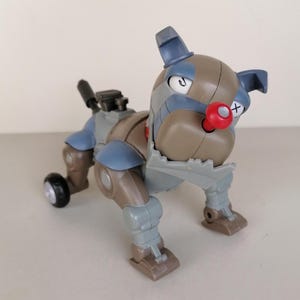Pode incluir: Um brinquedo de cão robótico em tons de cinza, marrom e azul, com um nariz vermelho e uma roda preta. O brinquedo tem um design mecânico com juntas visíveis e uma aparência estilizada.