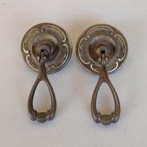 May include: A pair of antique brass drawer pulls. Each pull has a circular backplate with an ornate floral pattern and a teardrop-shaped handle. The pulls have a vintage, aged appearance.