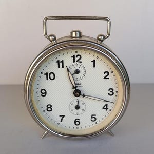 May include: A vintage silver-toned alarm clock with a white face and black numerals. The clock has a handle at the top and two small dials. The clock face has the word "SLAVIA" printed on it.
