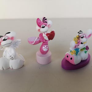 Könnte beinhalten: Drei skurrile Figuren von Zeichentrickfiguren. Eine ist weiß mit Engelsflügeln, eine andere ist rosa mit Ballettschuhen und einem Herzen, und die dritte ist weiß und rosa mit Blumen. Die Figuren stehen auf einer hellen Oberfläche.