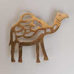 May include: A gold-coloured metal camel-shaped decorative object. The camel has an openwork design on its body and is standing on four legs. The head is detailed with an eye and a mouth. Likely a coaster or decorative piece.