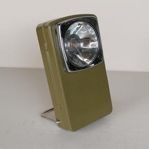 Può includere: Torcia vintage verde oliva con una lunetta cromata attorno alla luce. La torcia rettangolare ha un piccolo supporto in metallo. La luce è leggermente inclinata verso sinistra.