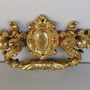 May include: A gold-toned ornate metal drawer pull with a decorative center design and two side handles.