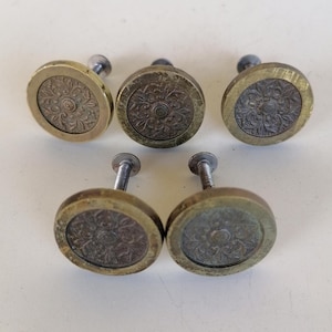 May include: Five antique brass cabinet knobs. Each knob features a circular design with a floral pattern in the center. The knobs have a tarnished, aged appearance, with a brass-colored rim and a darker, possibly bronze-colored center.