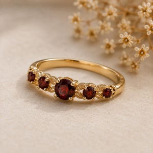 May include: A gold ring featuring five round, deep red garnet gemstones set in a row. The band is crafted with intricate, leaf-like metal detailing between each stone, with the largest garnet positioned in the center of the setting.