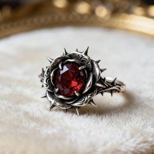 May include: A silver-toned ring featuring a rose design with thorns. The center of the rose holds a large, oval-cut, deep red gemstone. The ring's band also has thorn details, creating a gothic or fantasy aesthetic.