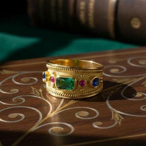 Peut inclure: Une bague dorée ornée d'une pierre verte centrale de taille émeraude, flanquée d'un rubis et d'un saphir. La bague a une bande texturée et est posée sur une surface en bois avec des motifs décoratifs. La bague est un bijou.