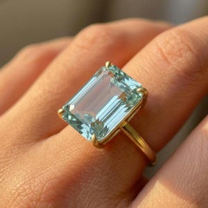 May include: A gold ring featuring a large, rectangular, light blue gemstone. The gemstone is set in a gold band with a simple, elegant design. The ring is shown on a person's finger.