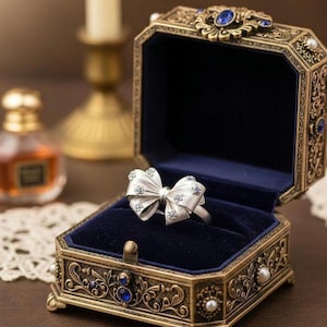 Puede incluir: Un anillo de plata con forma de lazo con pequeñas piedras preciosas, exhibido en un joyero ornamentado de estilo antiguo con interior de terciopelo azul. La caja es dorada con detalles intrincados y detalles azules.
