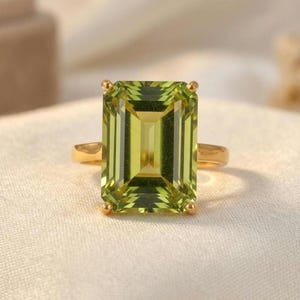 Può includere: Un anello d'oro con una grande pietra preziosa rettangolare verde chiaro. La gemma ha un design a taglio a gradini e la fascia dell'anello è in oro lucido. L'incastonatura ha quattro piccole griffe d'oro.