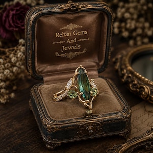 Può includere: Un anello in oro con una grande pietra verde taglio marquise, pietre verdi rotonde più piccole e pietre trasparenti, presentato in un cofanetto foderato in velluto marrone con il testo Rehan Gems And Jewels stampato in oro.