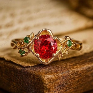 May include: A gold ring with a large, round, red gemstone at its centre. The ring has a floral design with green accent stones and detailed embellishments. The ring is displayed on a wooden surface.